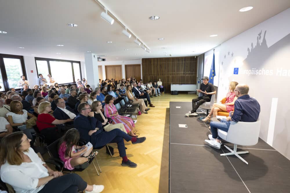 Auf dem Podium sitzen rechts 3 Personen im Gespräch. Links sitzen Schülerinnen und Schüler bei der Preisverleihung erliner Schülerwettbewerbs zur politisch-historischen Bildung 2025