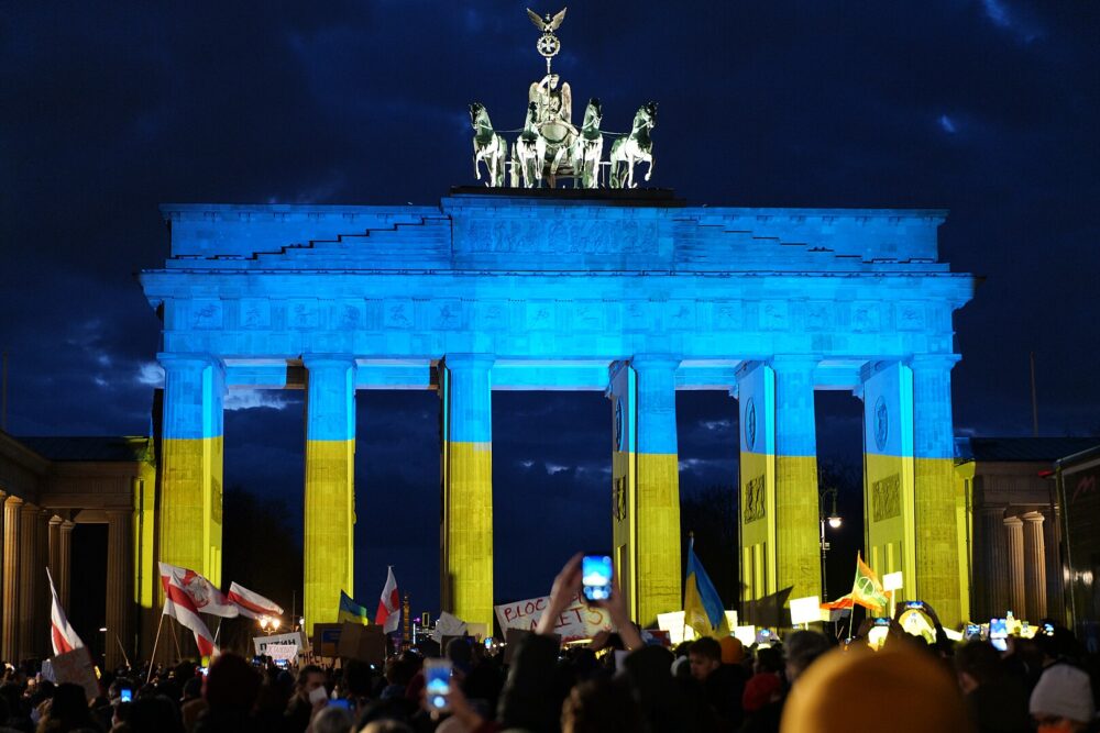 Das Brandenburger Tor am 24. Februar 2022, angestrahlt in den Nationalfarben der Ukraine. Im Vordergrund sind viele Menschen bei einer Solidaritätskundgebung für die Ukraine zu sehen,